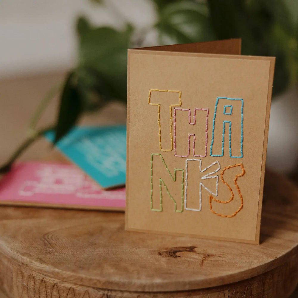 Card with 'THANKS' in colorful embroidery on a wooden surface with blurred greenery in the background