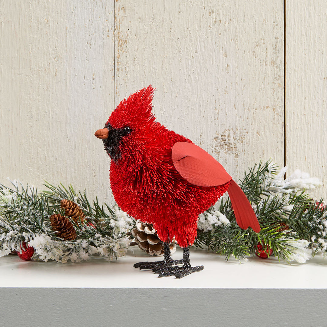 A red cardinal Christmas figure with textured red feathers, a black face, and a red wing, displayed on a white shelf with a snowy pine garland and pinecones against a rustic white wood background.