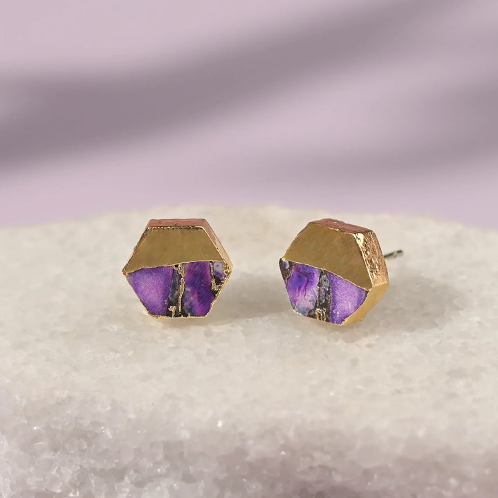 Hexagonal gold and purple earrings on a textured surface with a gradient background