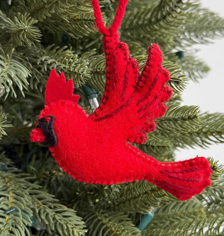 A bright red felt cardinal Christmas ornament hanging from a green pine tree branch, with a black beak detail and a red hanging loop.