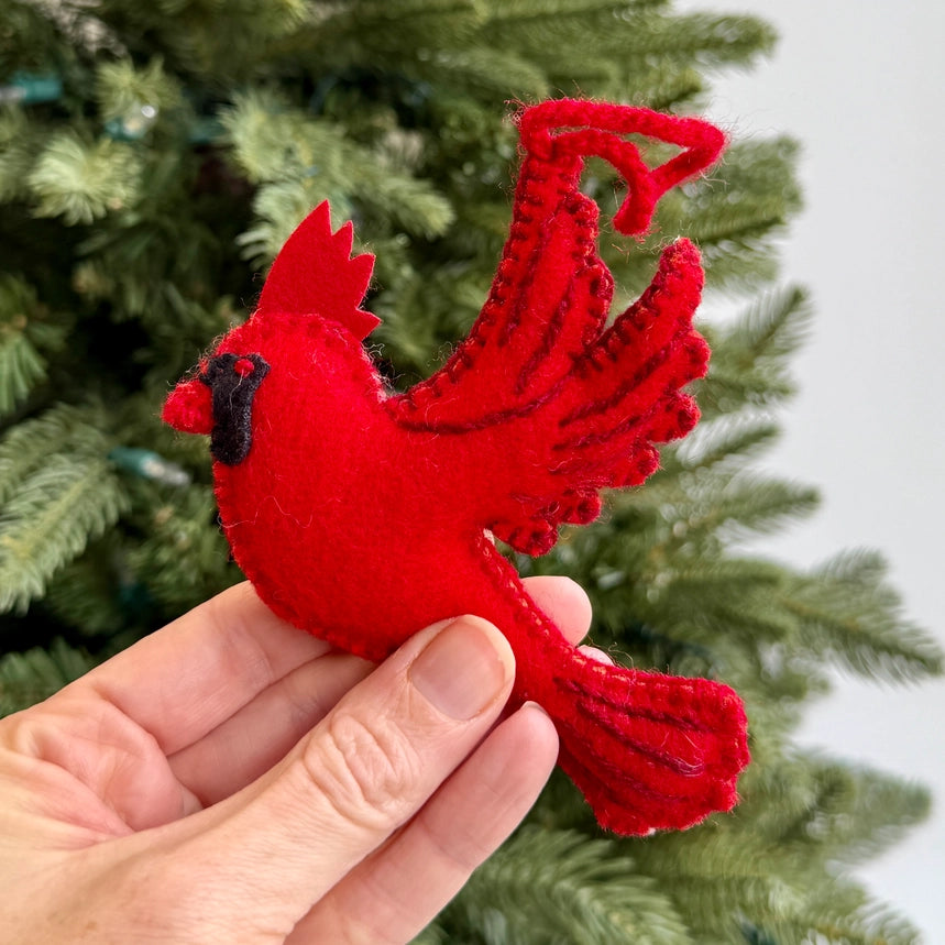 A hand holding a bright red felt cardinal Christmas ornament with black accents, set against a blurred green Christmas tree background.
