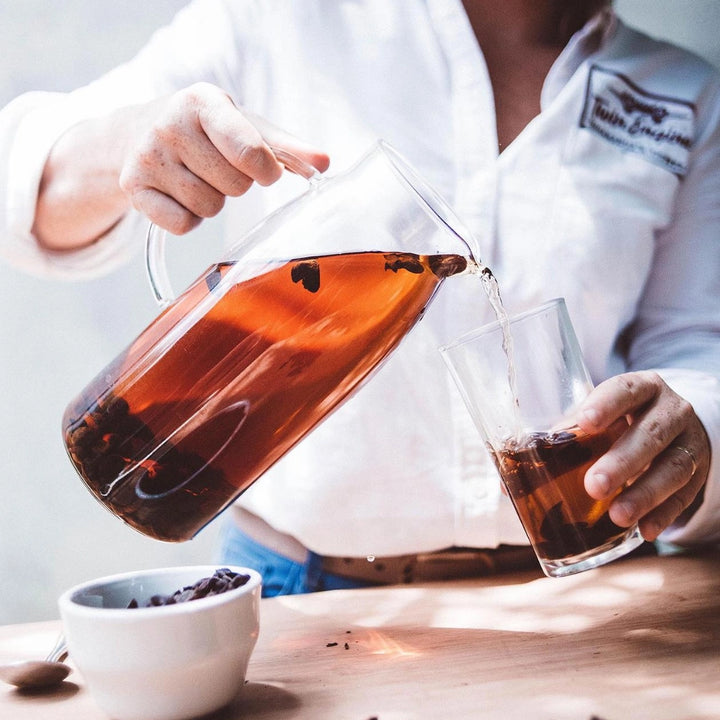 A person in a white shirt pours amber-brown tea from a clear glass pitcher into a clear glass tumbler, with a small white bowl on a wooden table in the foreground.