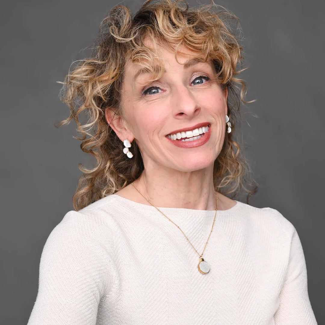 Woman with curly hair wearing a white sweater and gold necklace against a gray background