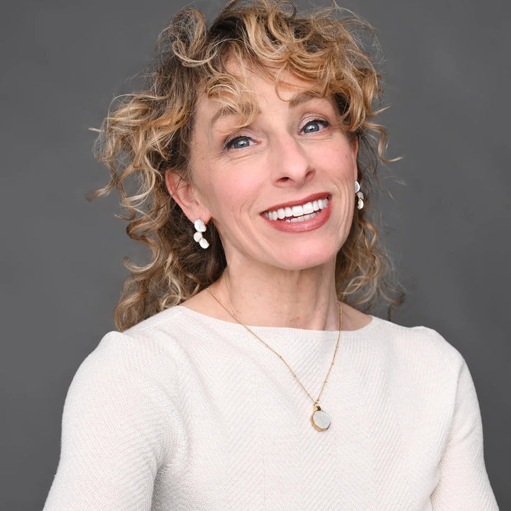 Woman with curly hair wearing a white sweater and gold necklace against a gray background