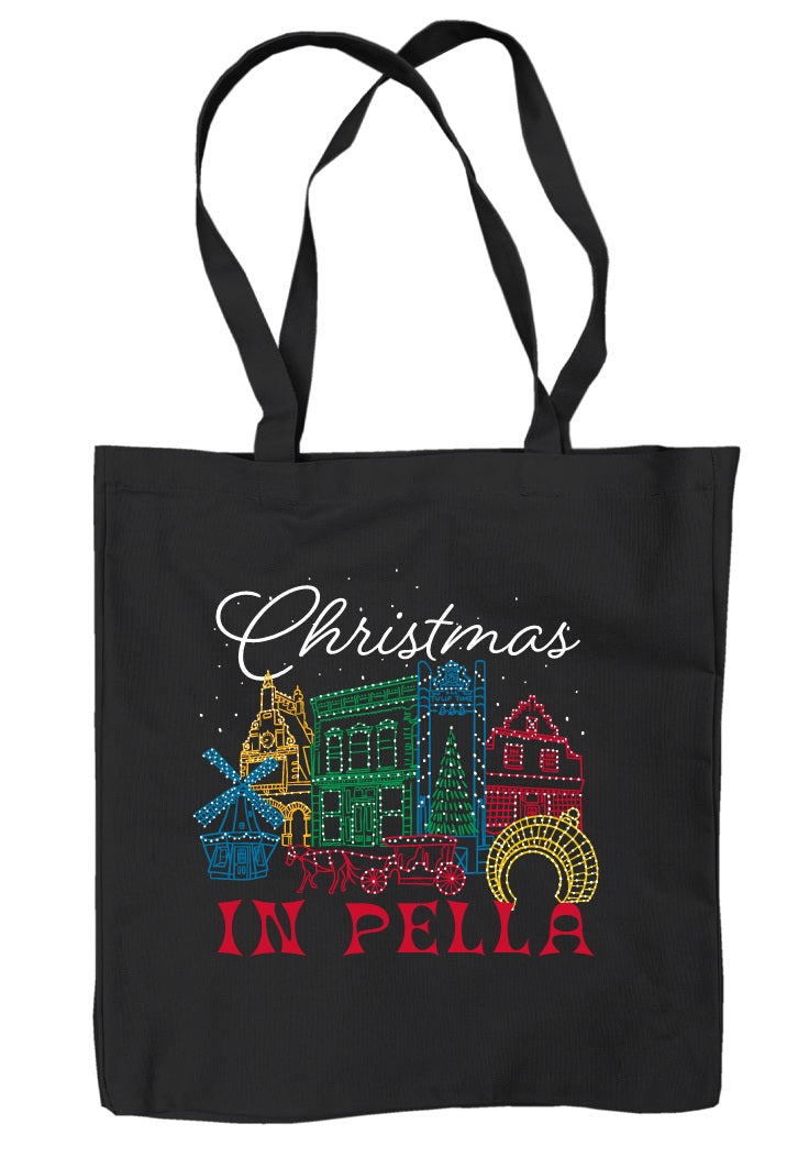 A black canvas tote bag with long black handles, featuring a colorful graphic of decorated Christmas buildings and the text 'Christmas IN PELLA' in white, green, blue, red, and yellow.
