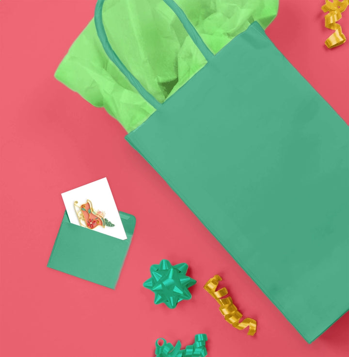 A flat-lay view of a green paper gift bag with a matching green tissue paper insert, set against a solid pink background with decorative gift bows and a small card.