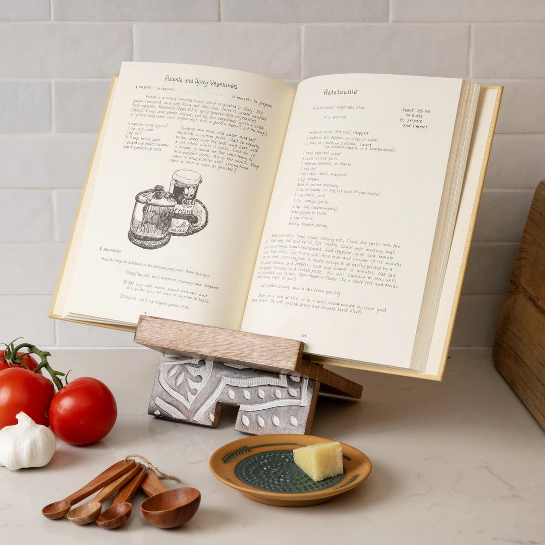 Open cookbook on a stand with tomatoes, garlic, and wooden spoons on a kitchen counter.