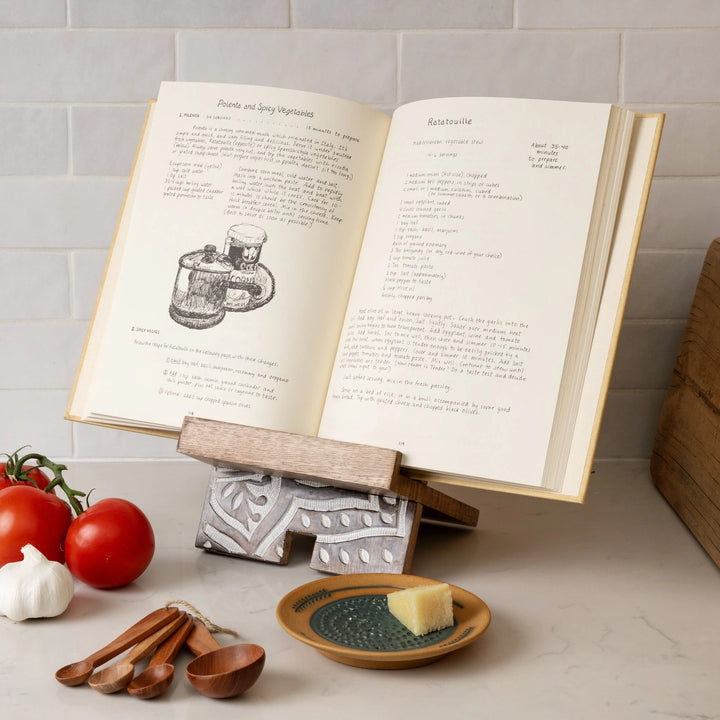 Open cookbook on a stand with tomatoes, garlic, and wooden spoons on a kitchen counter.