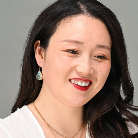 Woman with long dark hair wearing earrings and a necklace against a gray background