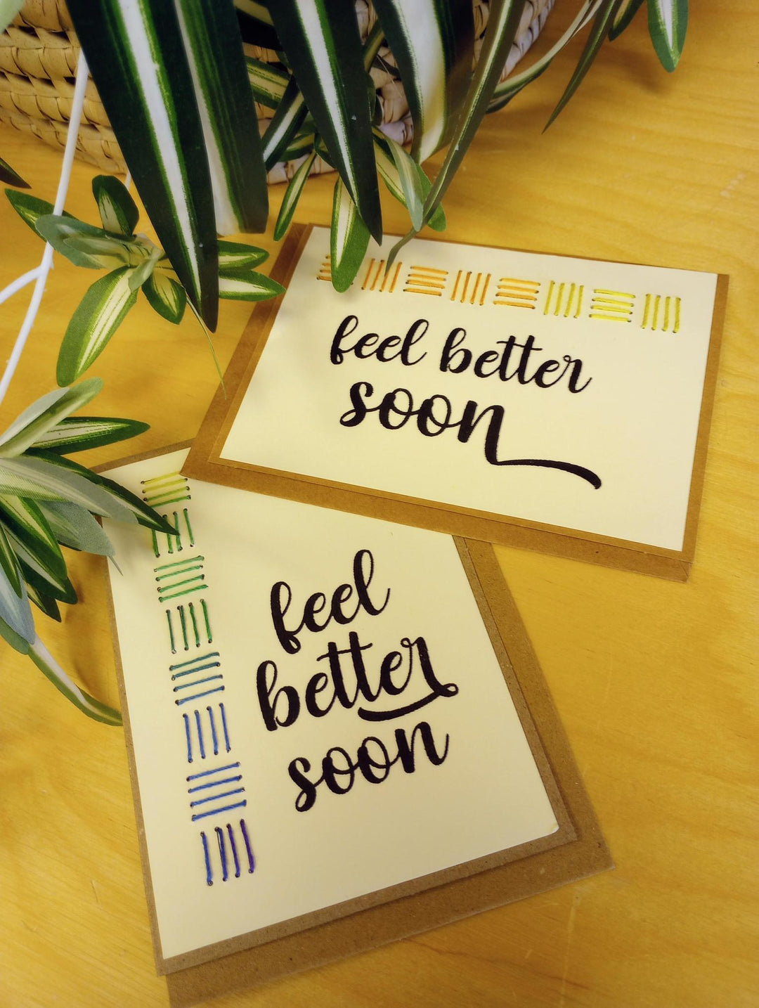 Two 'feel better soon' cards on a wooden surface with plants in the background