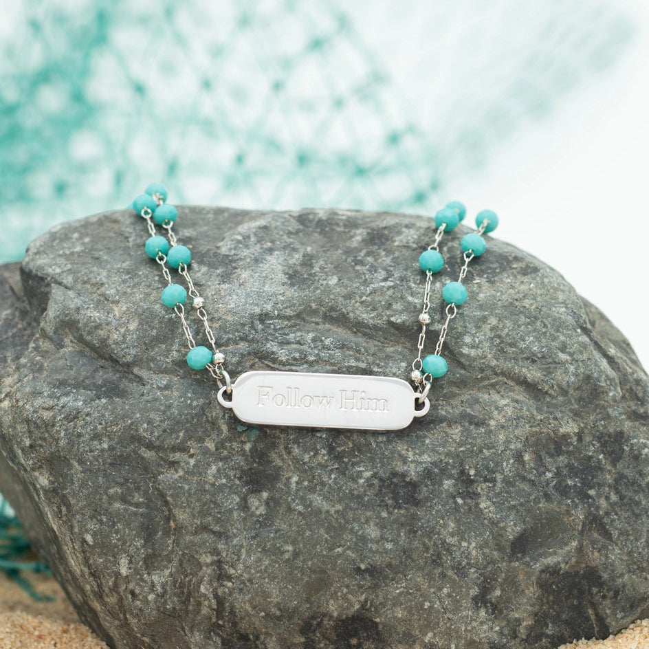 Turquoise beaded bracelet with a silver tag on a rock with a blurred green and white background