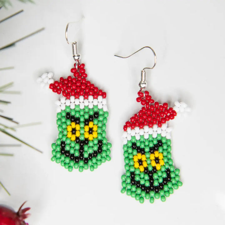 A pair of handmade beaded dangle earrings shaped like the Grinch, featuring green, red, white, yellow, and black seed beads, displayed on a white background with pine needles.
