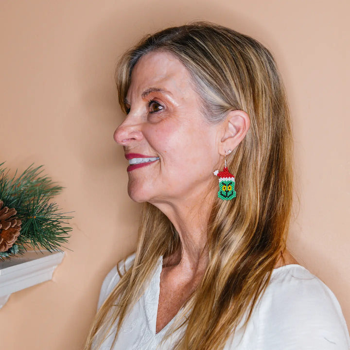 A woman with blonde hair is shown in profile wearing a colorful Grinch beaded earring. The earring features a green beaded lower section with a yellow center, a red beaded top, and a small white bead accent.
