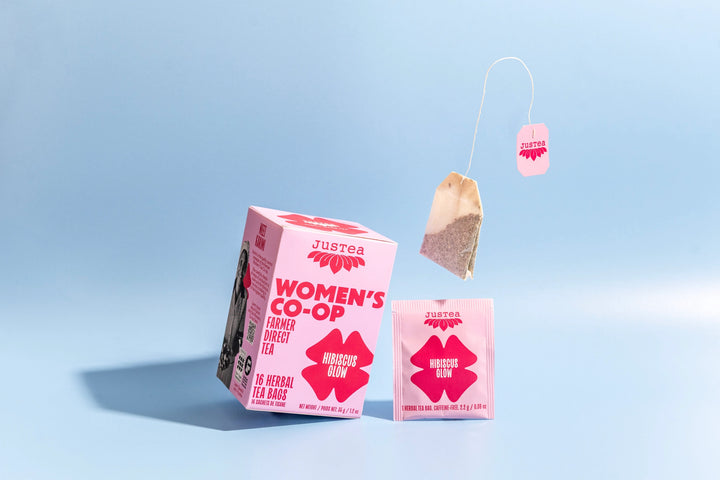 Justea Women's Co-op Hibiscus Glow tea boxes and a teabag on a light blue background