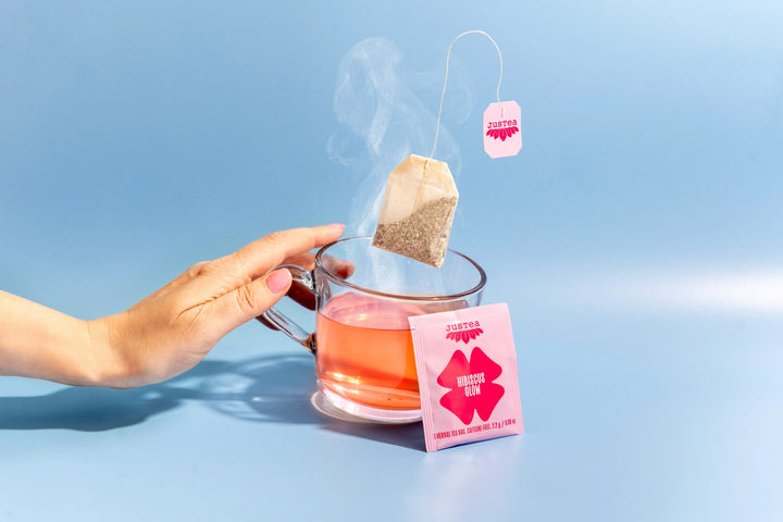 Hand holding a steaming cup of Hibiscus Glow tea with a tea bag on a blue background