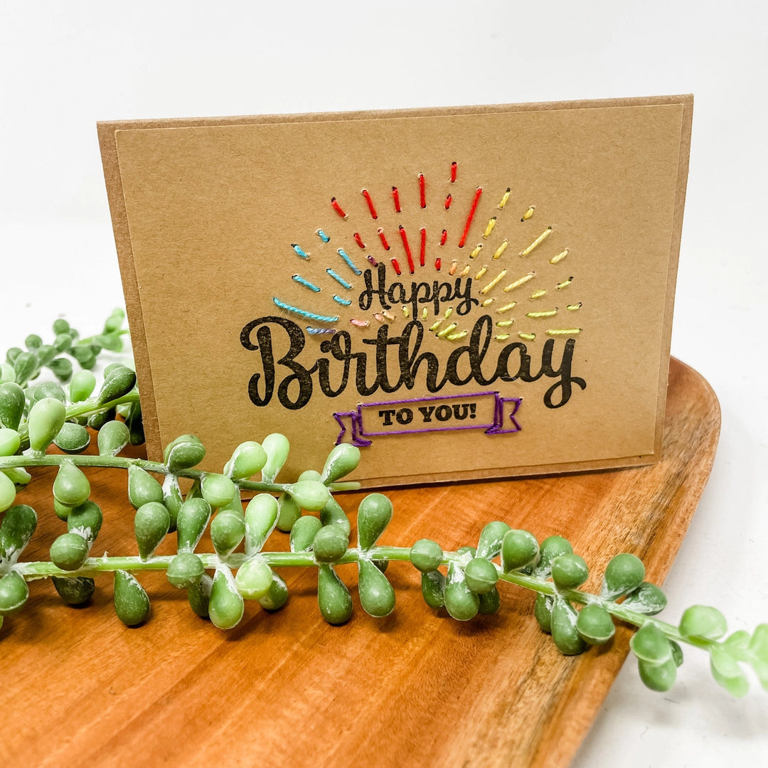 Birthday card with colorful text on a wooden surface with greenery