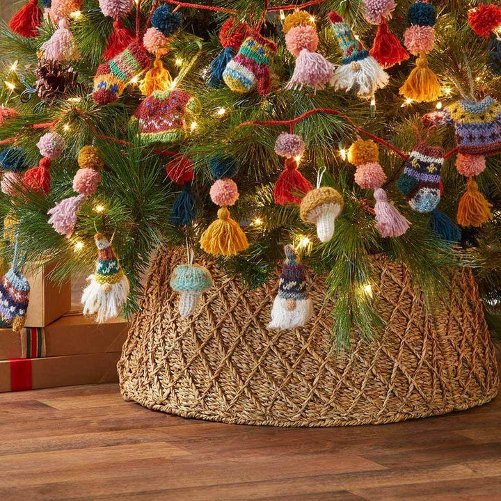 A woven round tree collar in natural holga fiber holding a decorated Christmas tree with colorful knitted ornaments, pom-poms, tassels, and warm string lights, with wrapped gifts and a wood floor visible.