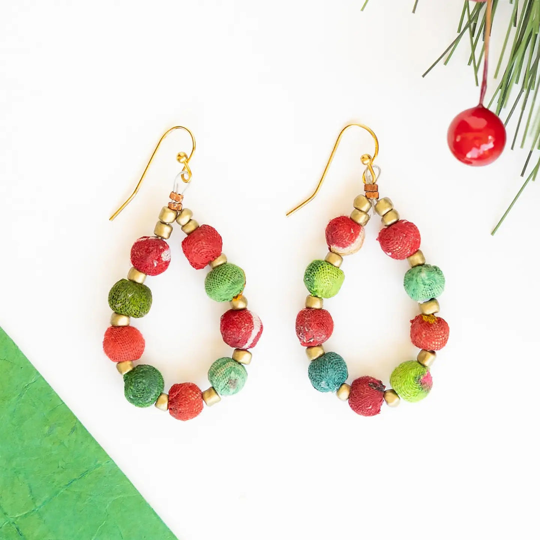 A pair of handmade dangle earrings with gold-tone fishhook wires and circular beads in red, and green, accented with small gold spacer beads. The earrings are displayed on a clean white background with decorative holiday greenery and a red berry in the top right corner.