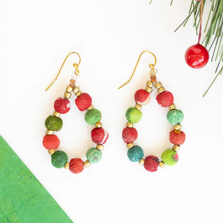 A pair of handmade dangle earrings with gold-tone fishhook wires and circular beads in red, and green, accented with small gold spacer beads. The earrings are displayed on a clean white background with decorative holiday greenery and a red berry in the top right corner.