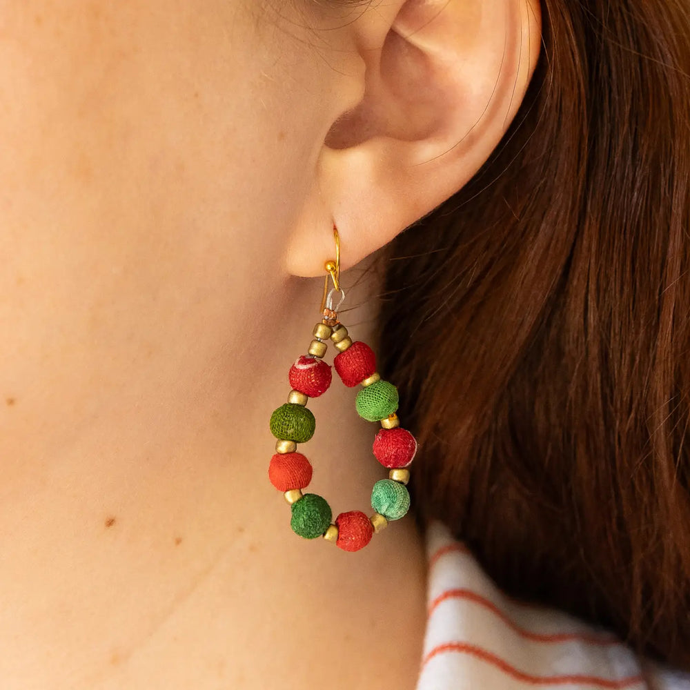 A close-up of a person's ear wearing a handmade hoop earring made of red and green textured beads with small gold spacer beads and a gold hook.