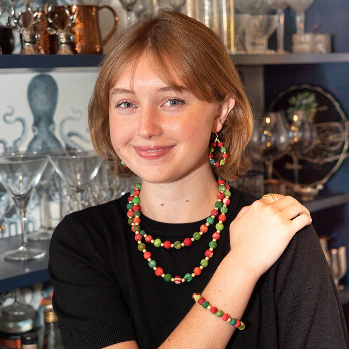 A woman with light brown hair wearing a colorful beaded necklace, matching drop earrings, and a beaded bracelet, posed in front of a bar shelf filled with glassware and copperware.