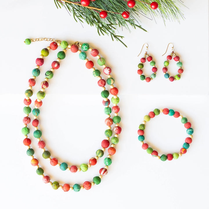 A coordinated jewelry set laid flat on a white background, including a long beaded necklace, matching drop earrings, and a beaded bracelet. The beads are marbled in red and green with small gold-tone metal findings.