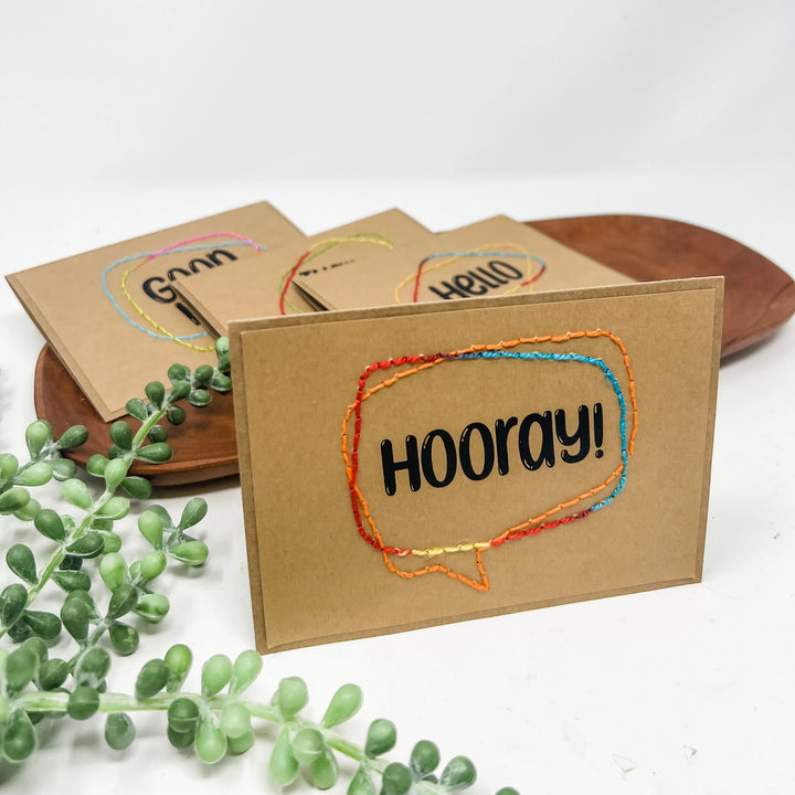 Kraft paper card with 'HOORAY!' text and colorful border on a white background with greenery.