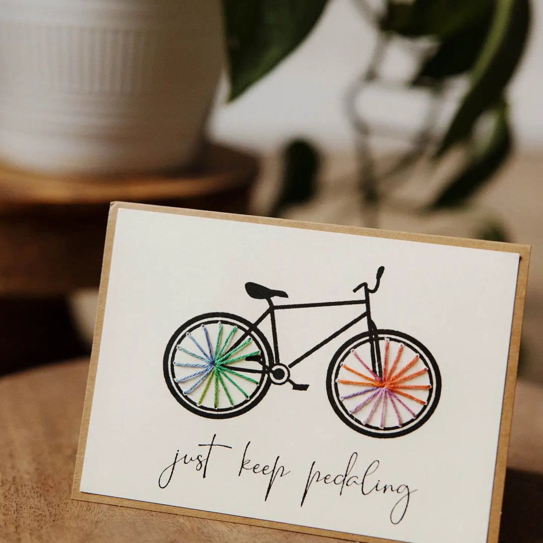 Card with a bicycle illustration and 'just keep pedaling' text on a wooden surface.