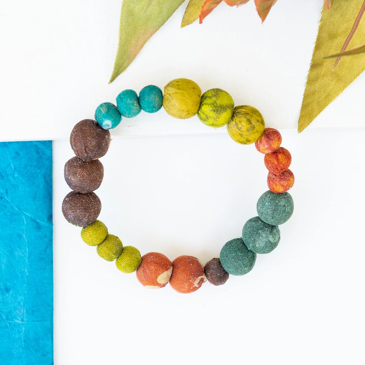 A handmade beaded bracelet made of round, earthy-colored wooden beads in a rainbow sequence, arranged in a circle on a white background with decorative leaves and a blue textured strip.