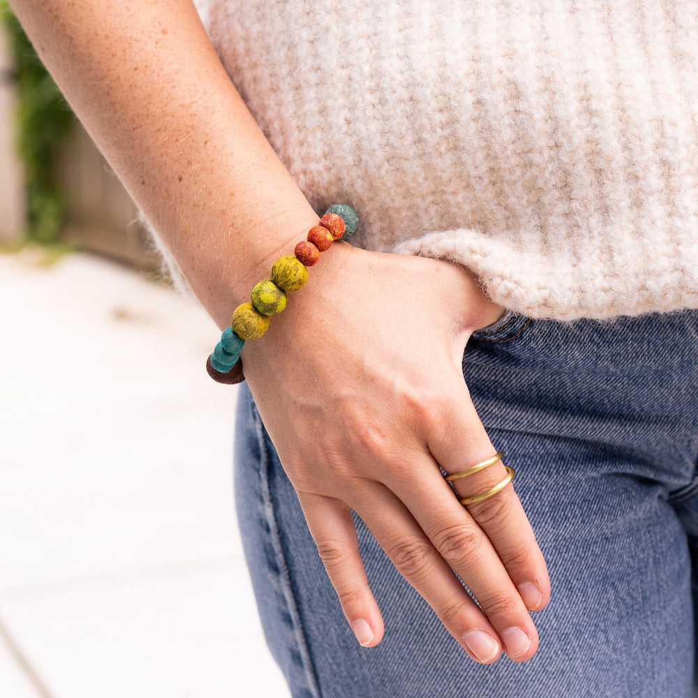 A close-up of a person's hand and wrist wearing a colorful beaded bracelet. The bracelet features round beads in teal, olive green, mustard yellow, terracotta orange, and dark brown.