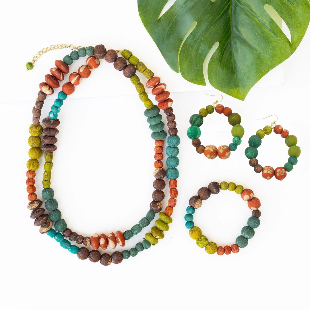 A multi-piece beaded jewelry set featuring a long necklace, two hoop earrings, and a bracelet, all made with round beads in earthy brown, teal, orange, and olive green. The set is styled on a white background with a large green monstera leaf in the corner.