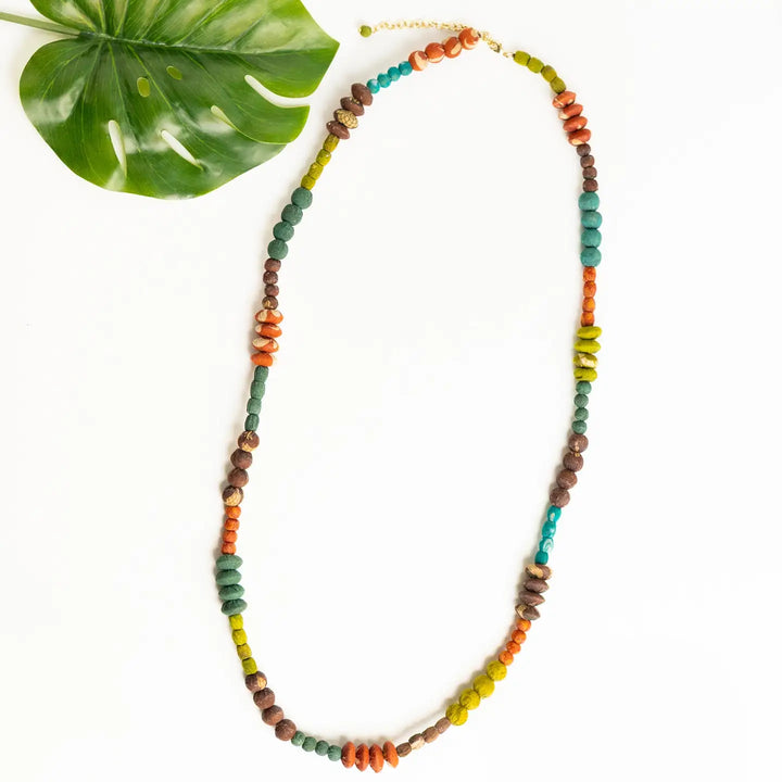 A handmade beaded necklace with multiple colors of wooden beads (green, brown, orange, yellow, teal) arranged in a long loop on a white background, styled with a green monstera leaf in the top left corner.
