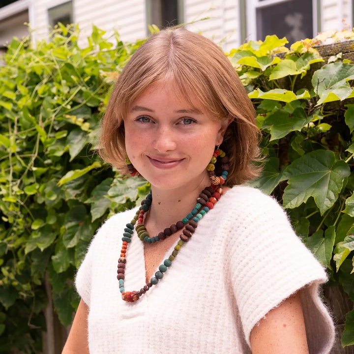A smiling woman with shoulder-length blonde hair wearing a white knit top and colorful beaded necklaces and earrings, standing outdoors in front of green leafy plants.