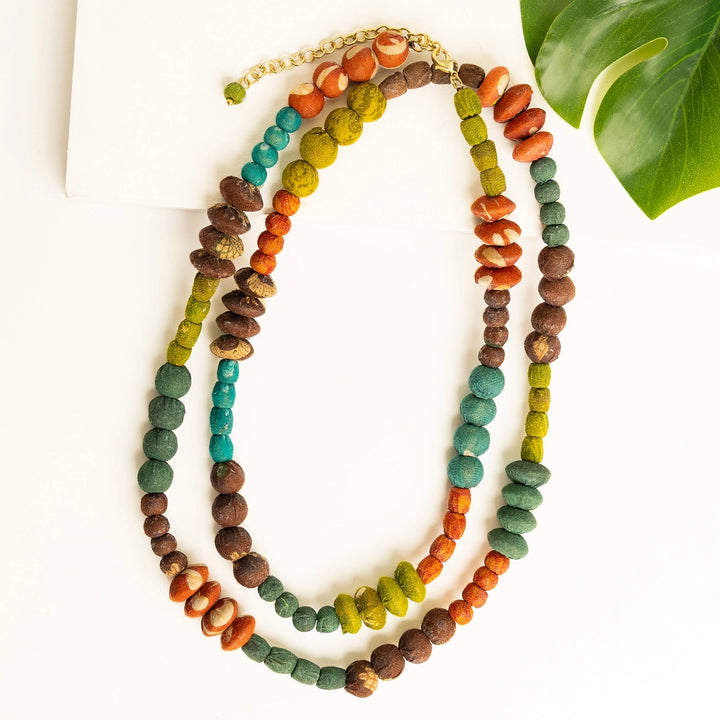 A multi-strand beaded necklace featuring natural wood beads in brown, turquoise, green, and orange, arranged on a white background with a green leaf prop.