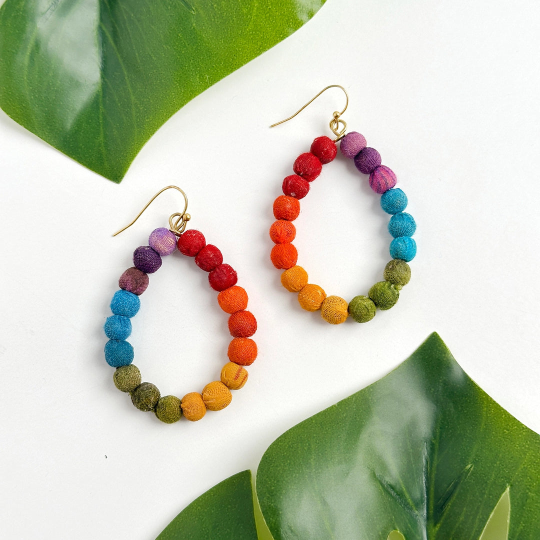 Colorful beaded Kantha teardrop hoop earrings on a white background with green leaves.