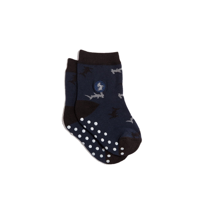 A pair of navy blue baby socks with grip dots on the sole, dark brown heels and cuffs, and a pattern of small grey sharks. A circular blue and white embroidered logo is visible on the side.