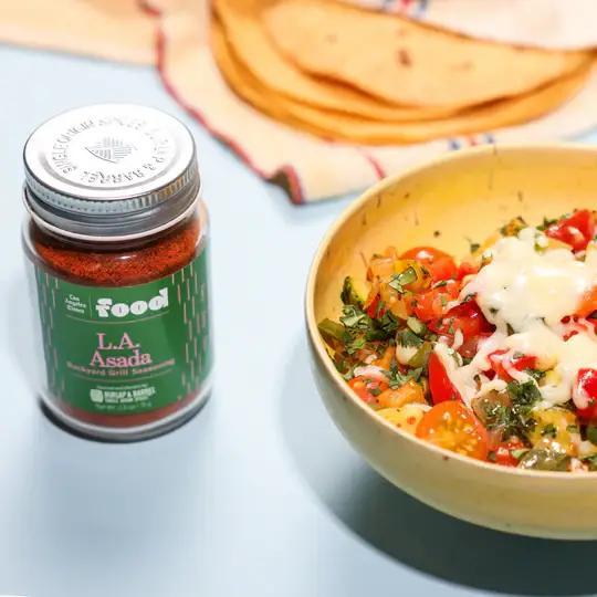 Jar of 'LA Asada' seasoning next to a bowl of salad and tortillas on a blue surface