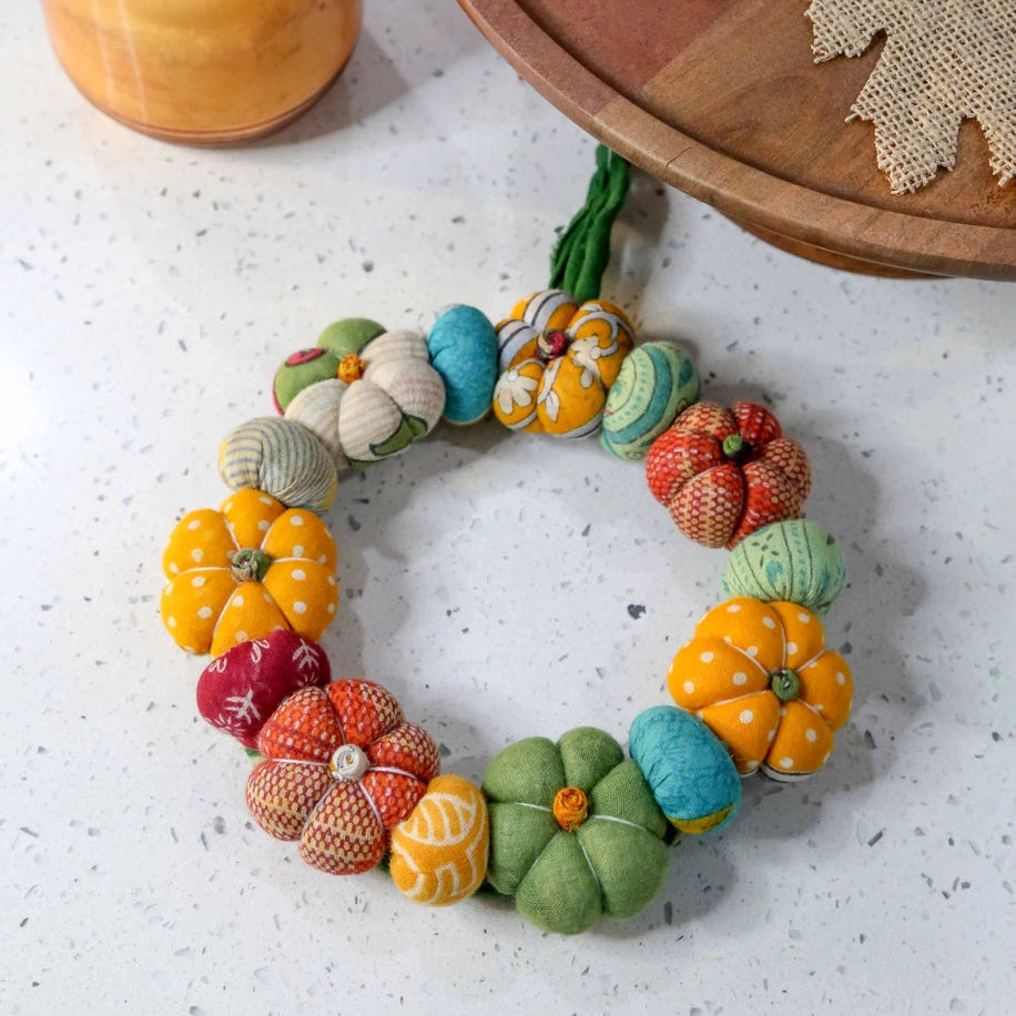 Colorful felt pumpkin wreath on a textured surface with a wooden tray and jar in the background.