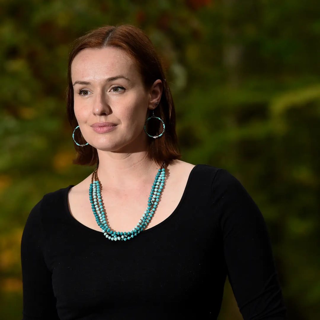 Woman wearing a black top and turquoise paper bead necklace with a blurred green background