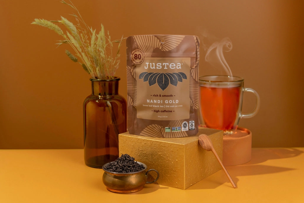 Justea Nandi Gold coffee package with a cup of tea and tea leaves on a warm-toned background
