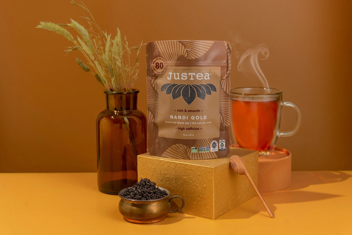 Justea Nandi Gold coffee package with a cup of tea and tea leaves on a warm-toned background