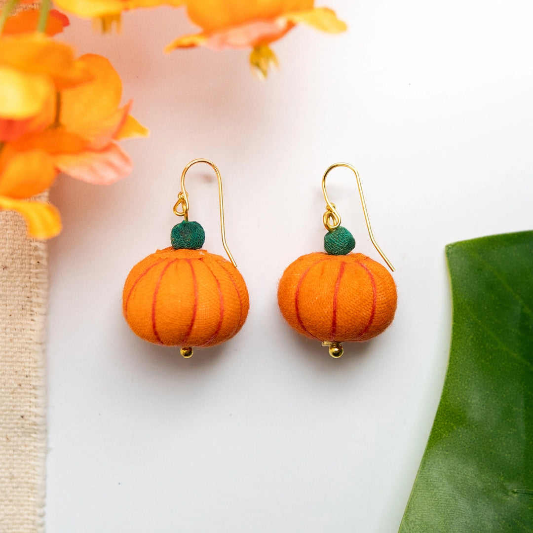 Pumpkin-shaped earrings with gold hooks on a white background with orange flowers and green leaves.