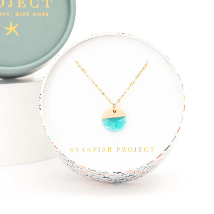 A gold necklace with a round aqua pendant in a Starfish Project gift box.