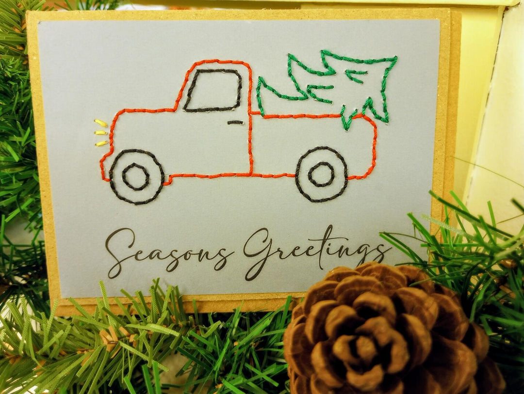 Embroidered card with a truck and tree design and 'Seasons Greetings' text, surrounded by greenery and a pine cone.