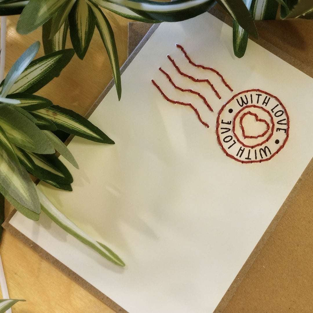 A square white greeting card with a red stamped postmark design reading 'WITH LOVE' around a heart, photographed on a tan surface with green leaves around the edges.