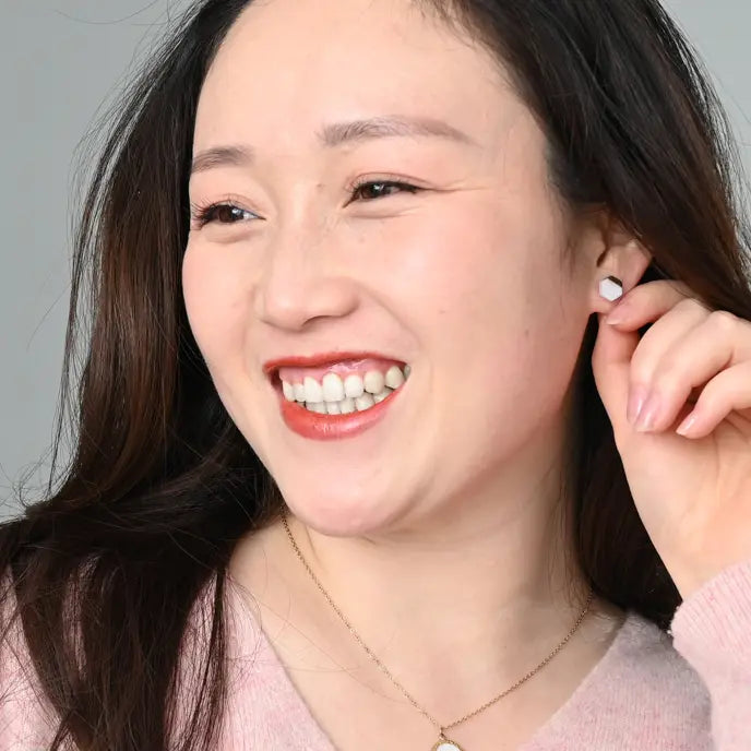 A close-up portrait of a smiling woman with dark hair, wearing a small white stud earring and a delicate gold necklace, set against a plain light gray background.