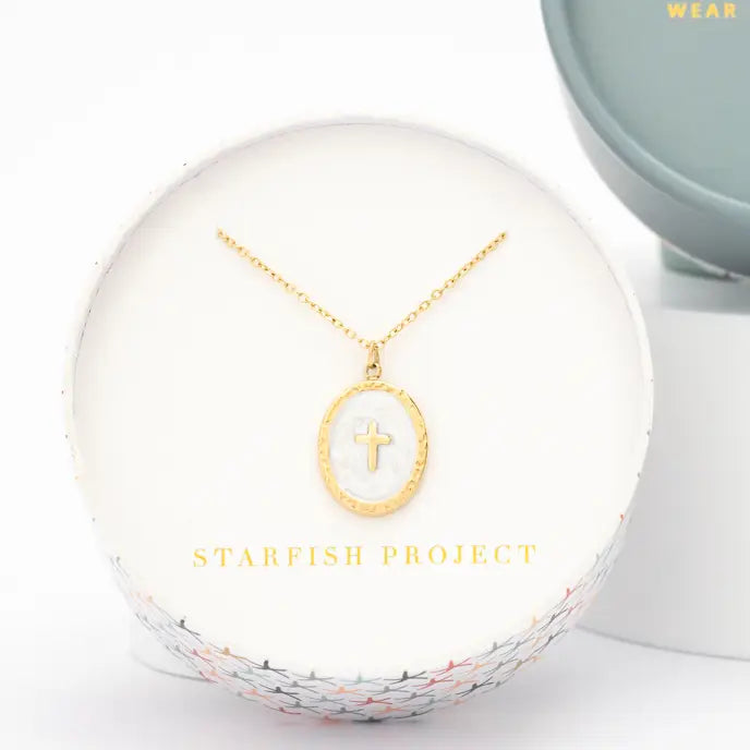 A gold necklace with an oval pendant featuring a small cross, displayed on a white circular presentation box with the brand name 'STARFISH PROJECT' printed in gold. A small blue jewelry stand is partially visible in the background.