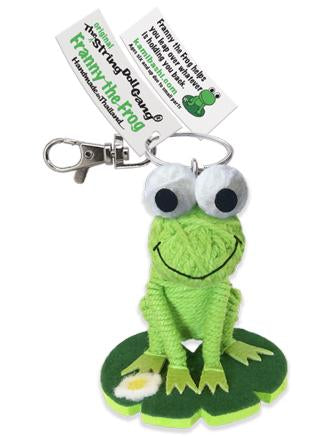 A green frog string doll keychain with large white and black eyes, a stitched smile, and a green lily pad base, attached to a metal clasp with a branded paper tag.
