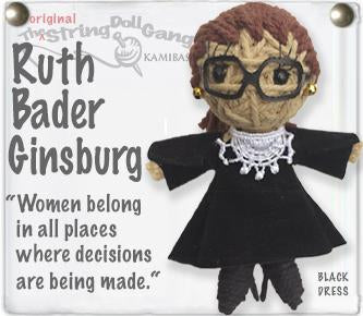 A handmade string doll of Ruth Bader Ginsburg with brown yarn hair, black glasses, a black dress, and a white lace collar, mounted on a white display card with text.