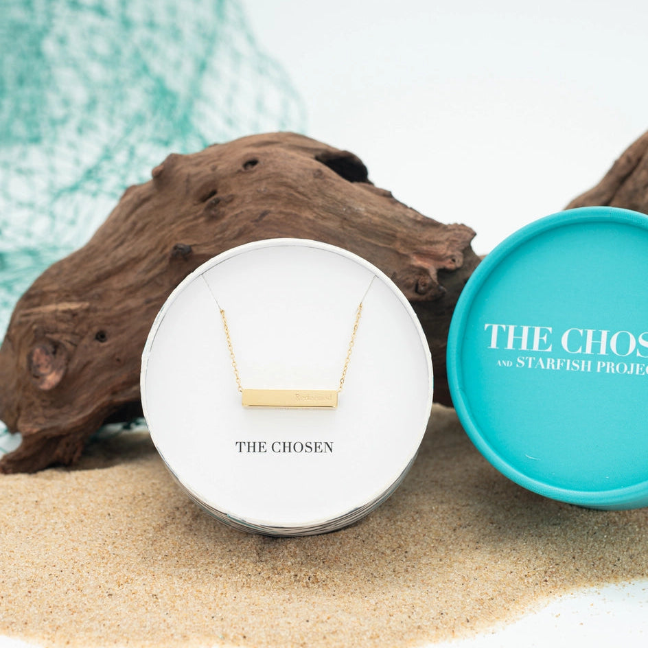Gold necklace in a white display box with 'The Chosen' branding, placed on sand with starfish and driftwood.